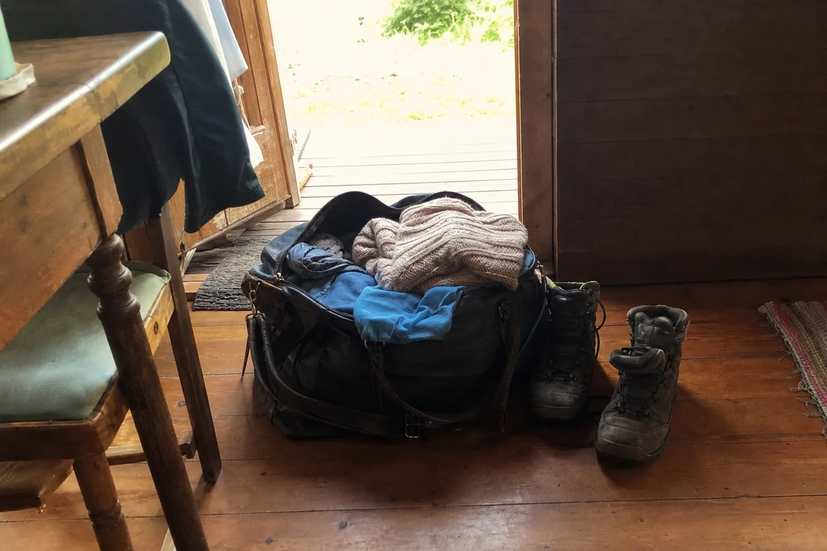 A packed weekend bag with warm layers and boots by a cabin door, lived-in, unstaged