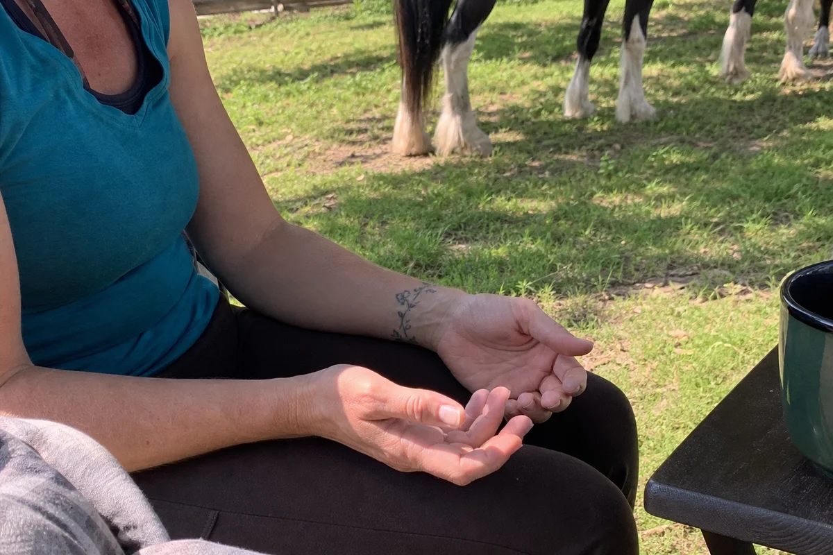 Detail of hands resting open on a lap outdoors, calm, with a horse's legs softly out of focus behind