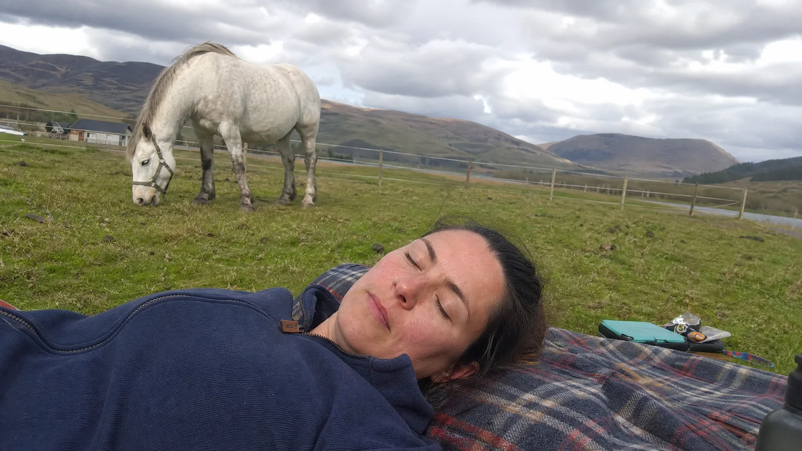 Person resting on a blanket in a paddock, eyes closed, a horse grazing nearby, soft overcast Highlands light
