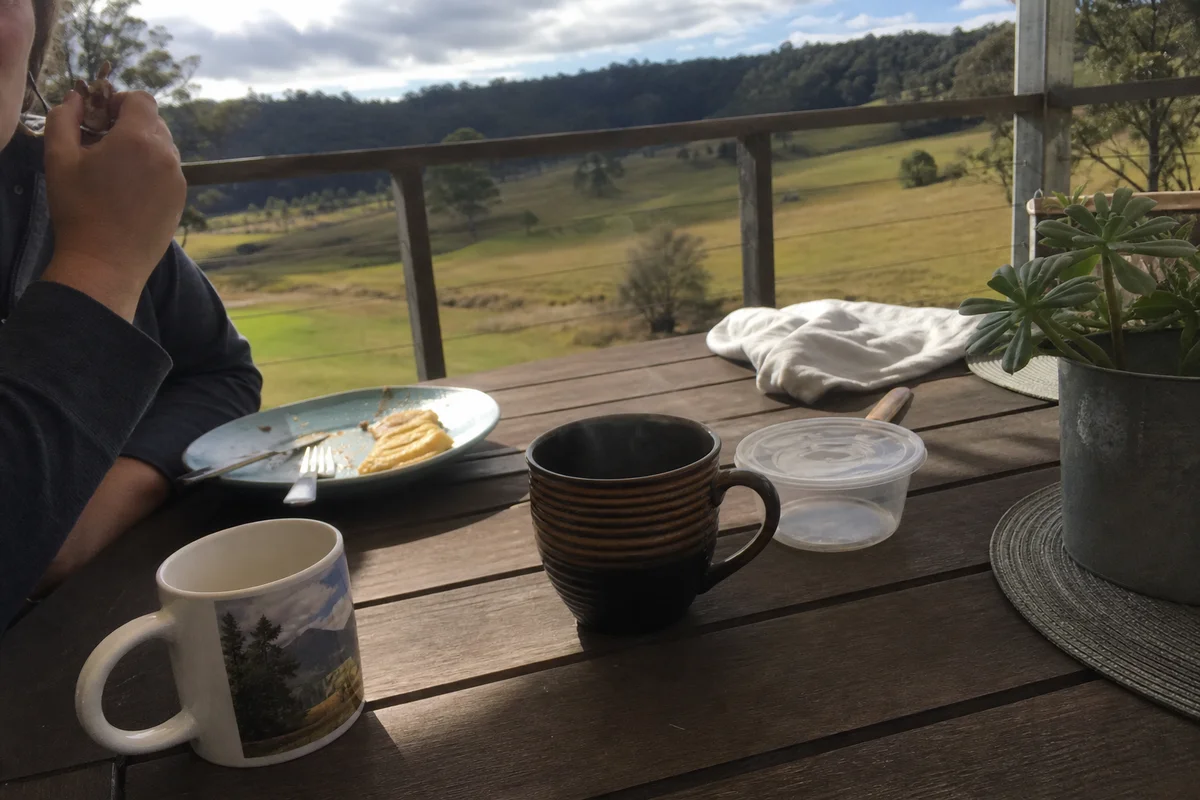 Simple breakfast on a timber deck overlooking paddocks, steam from a mug, lived-in and calm