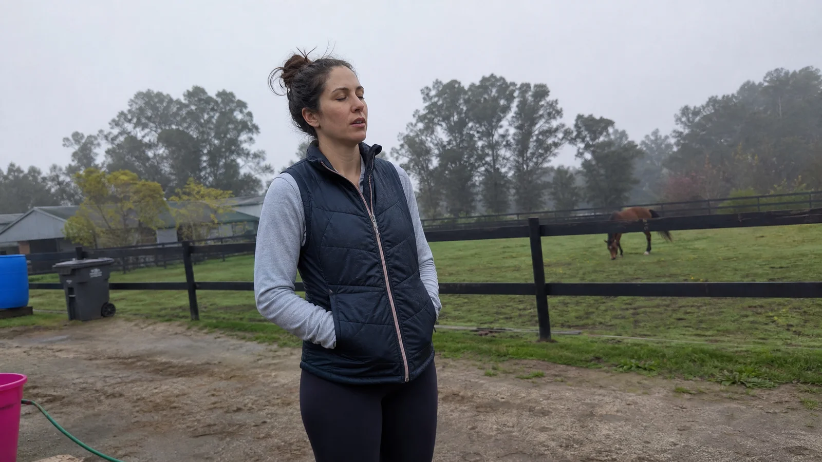 Woman exhaling slowly, shoulders dropping, standing in a quiet paddock with a horse grazing behind her, cool morning