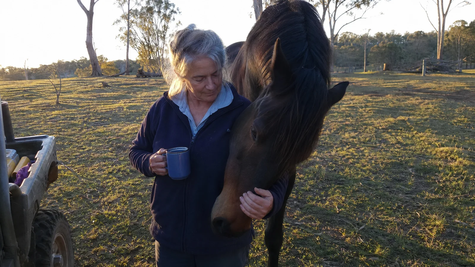 Woman in her 50s standing peacefully in a paddock at golden hour, a horse's head lowered near her shoulder